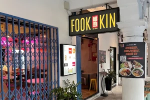 The exterior entrance of Fook Kin restaurant featuring a modern black and yellow sign and traditional blue folding gates.