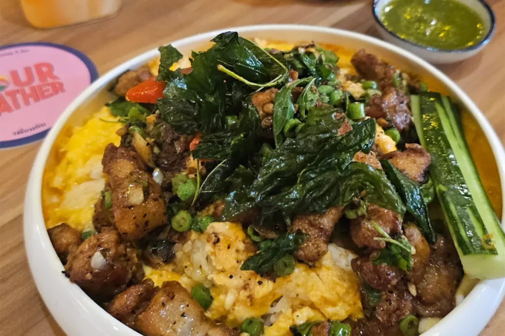 A close-up of a Thai basil pork rice bowl topped with crispy fried basil leaves, served with a side of cucumber and green chili sauce.