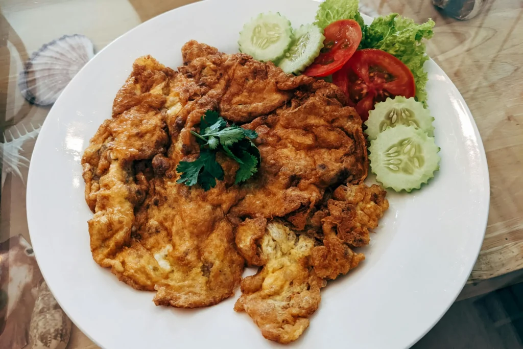A golden-brown, crispy Thai-style omelet sits on a white plate, garnished with a sprig of fresh cilantro. It is served alongside a side of sliced cucumbers, tomatoes, and green lettuce leaves on a wooden table.