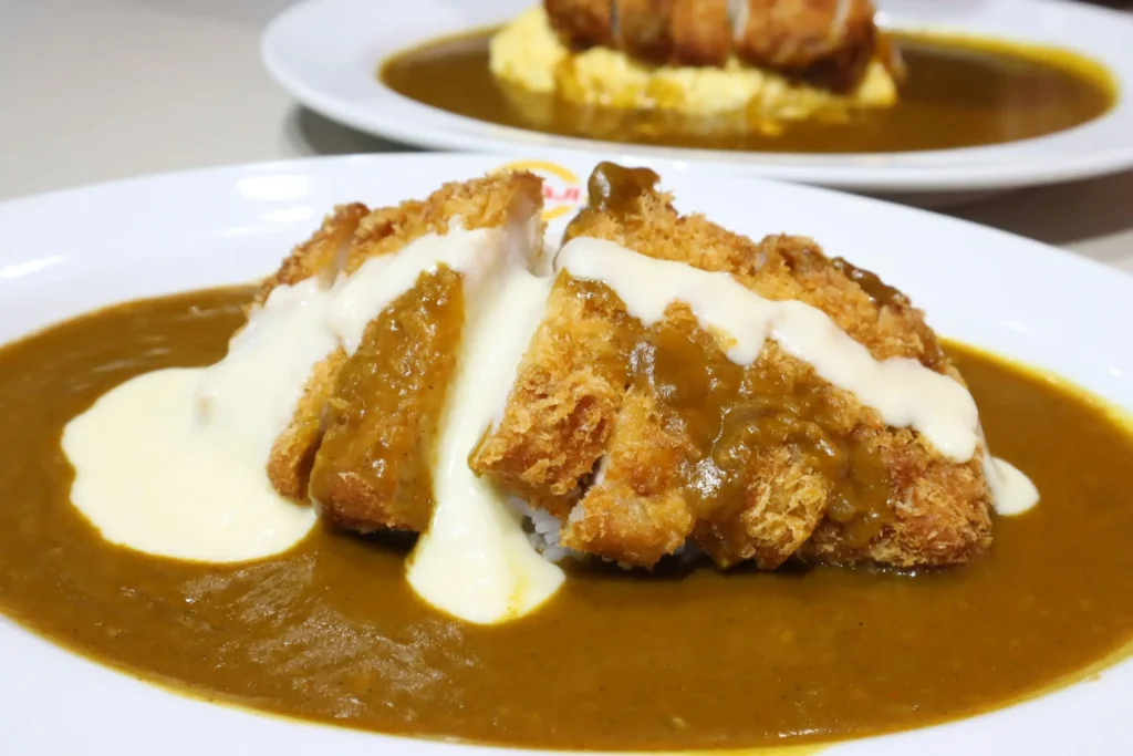 This image features a plate of Japanese curry topped with a breaded cutlet and a generous drizzle of white cheese sauce. In the background, a second plate shows a similar dish served over a bed of fluffy omelet rice.