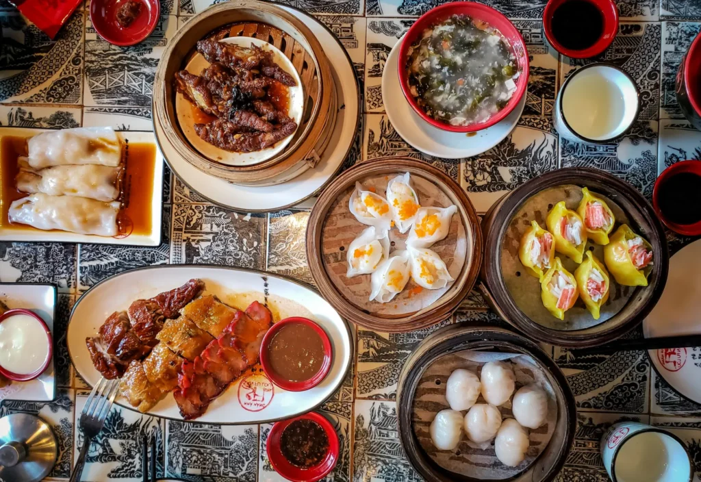 This top-down photograph captures a vibrant spread of dim sum and Cantonese dishes arranged on a patterned tile tabletop. The assortment includes bamboo steamers of dumplings, plates of roasted meats, and bowls of soup, all presented with authentic textures and rich colors.