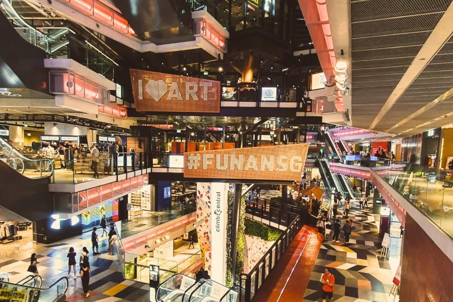 The image showcases the vibrant, multi-level interior of Funan Mall in Singapore, featuring an industrial-chic design with neon lighting and geometric floor patterns. Large glowing signs for "#FUNANSG" and "I Love ART" hang over the central atrium, while shoppers navigate the escalators and walkways surrounding a prominent climbing wall.