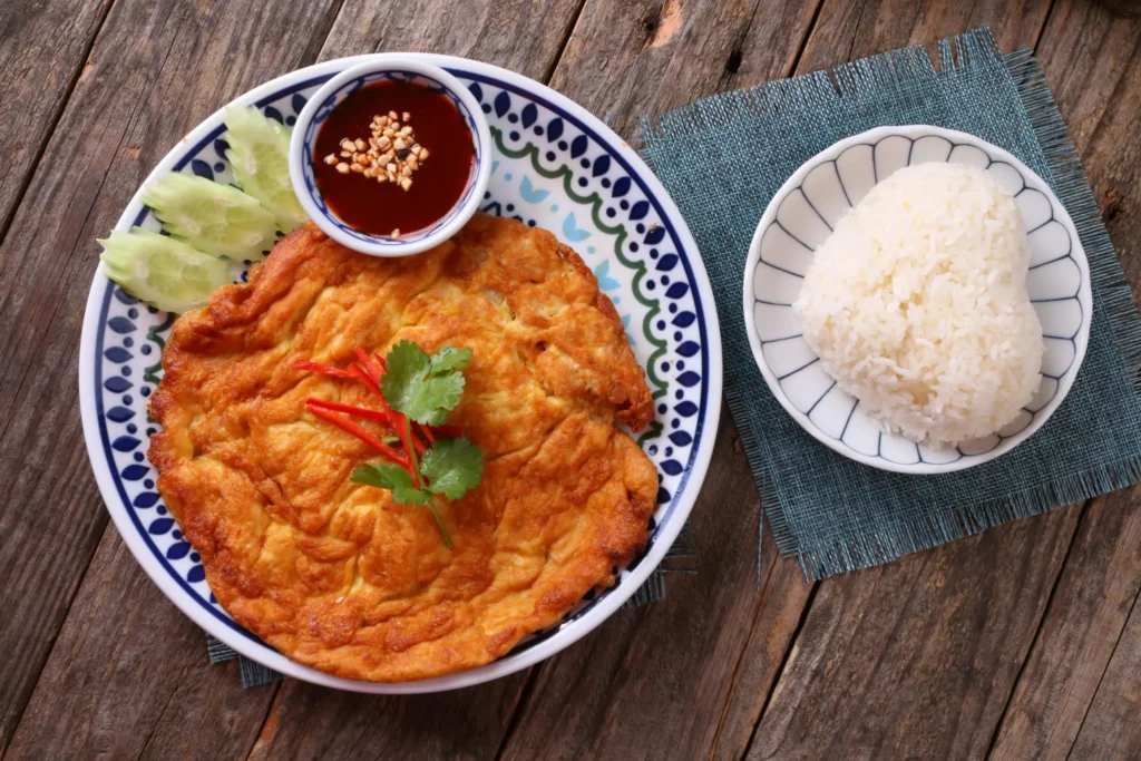 A golden-brown Thai-style omelet garnished with herbs and peppers is served on a decorative plate alongside sliced cucumbers and a small bowl of dipping sauce. A heart-shaped mound of white rice sits on a separate small plate to the right, all arranged on a rustic wooden tabletop.