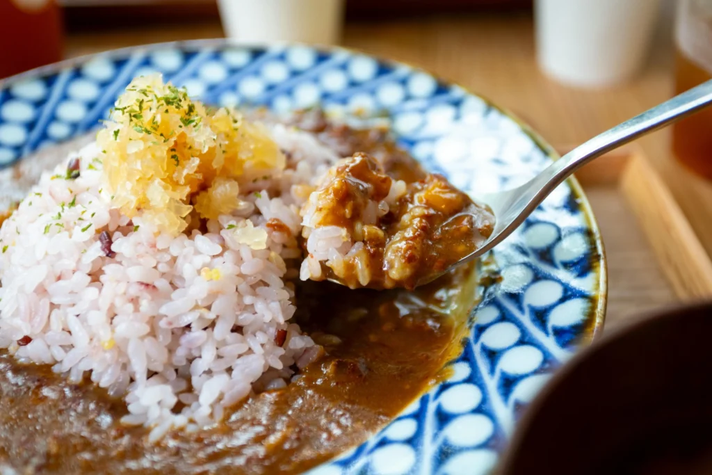A spoonful of Japanese curry is lifted from a plate piled with multi-grain rice and topped with pickled garnishes. The dish is served on a blue and white patterned ceramic plate, highlighting the rich, thick texture of the sauce.