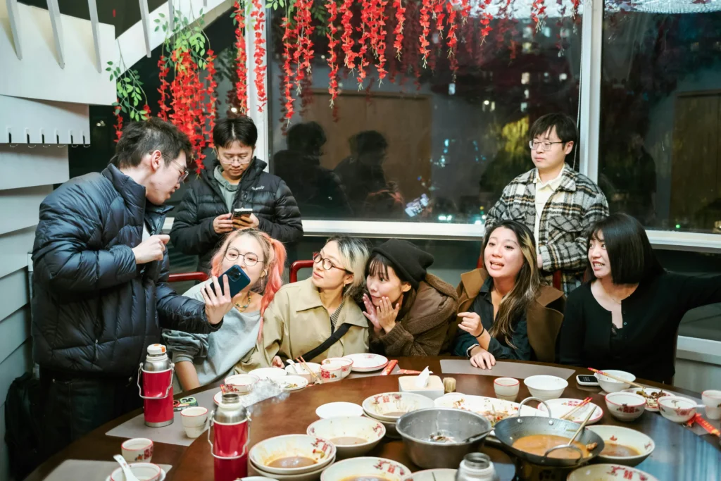A group of friends gathers around a circular dining table filled with finished bowls and plates after a meal. Several people lean in toward a man on the left who is holding up his phone to show them something on the screen.