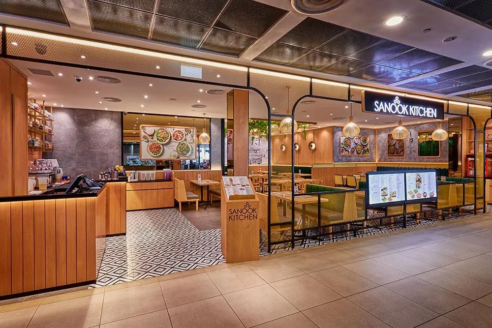 The image shows the warm and modern storefront of Sanook Kitchen at Parkway Parade, featuring light wood paneling and mesh partitions. The interior is brightly lit with birdcage-style pendant lamps over comfortable green and yellow seating.