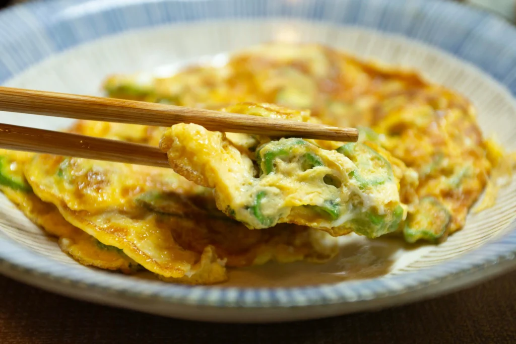 A pair of wooden chopsticks lifts a bite of a golden-brown omelet filled with sliced green vegetables. The dish is served in a blue-rimmed ceramic bowl, showcasing the fluffy texture of the fried eggs.