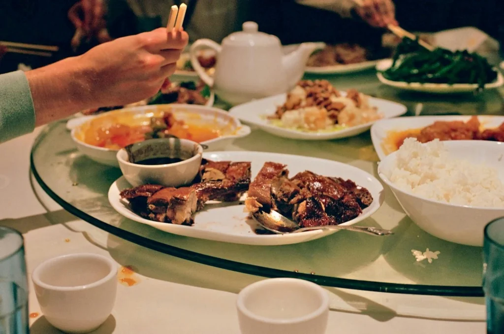 A variety of Chinese dishes, including roasted duck and white rice, are spread across a large spinning glass tray on a dining table. Several people are gathered around the meal, with one person using chopsticks to reach for a dish near a white teapot.