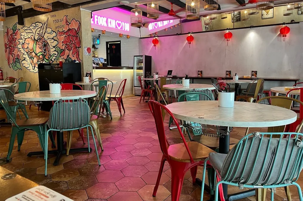 The interior of Fook Kin restaurant showing marble-topped tables, colorful metal chairs, and neon pink lighting with birdcage decor