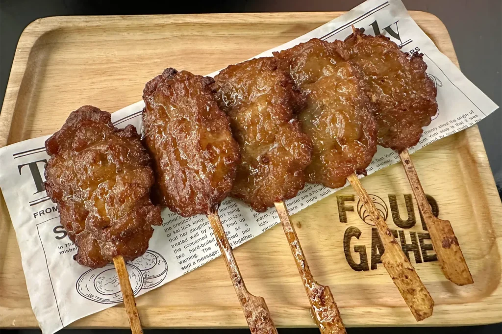 Five grilled Thai pork skewers (Moo Ping) served on a wooden tray lined with newspaper-themed parchment paper.