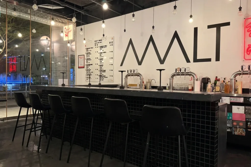 The sleek interior of MALT bar featuring a black tiled counter, multiple silver beer taps, and a large craft beer menu on the wall