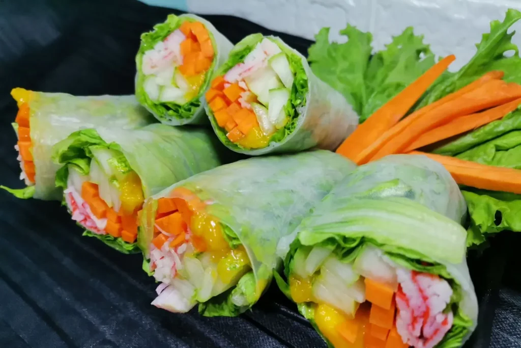 These fresh spring rolls are tightly wrapped in translucent rice paper, revealing vibrant layers of lettuce, julienned carrots, cucumber, and crab sticks. They are neatly arranged on a dark surface and garnished with extra leafy greens and carrot slivers.