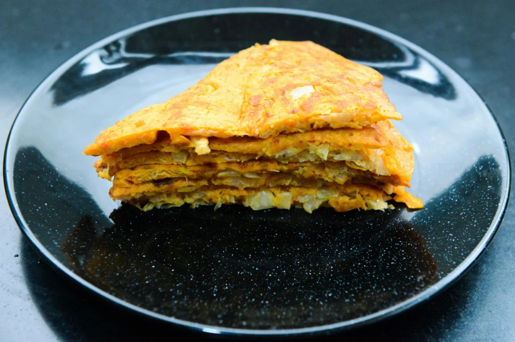 A stack of several triangular slices of orange-colored flatbread or savory pancakes sits on a black plate. The food appears to have a textured, layered interior filled with small pieces of chopped vegetables like onions or cabbage.