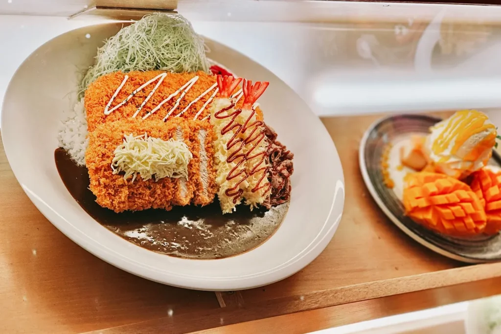 This image displays a large, hearty plate of Japanese-style curry topped with breaded cutlets, fried shrimp, and shredded cabbage. Beside it sits a smaller dish featuring fresh, sliced mango topped with a drizzle of sauce and a scoop of cream.