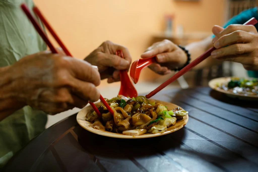 Two people sit at a dark table, using red chopsticks and spoons to share a plate of stir-fried flat rice noodles. The dish is tossed with green vegetables and a dark, savory sauce in a casual dining setting.