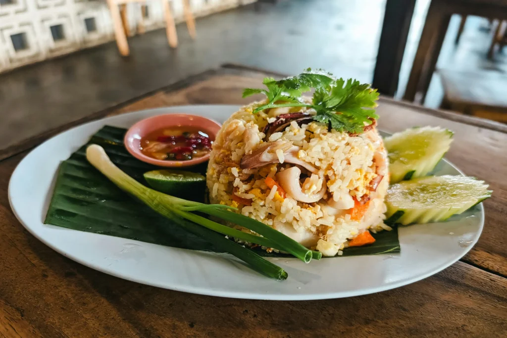 This image features a mound of seafood fried rice served on a banana leaf atop a white plate, garnished with fresh cilantro and pieces of squid. It is accompanied by classic sides including sliced cucumbers, a scallion, a lime wedge, and a small dish of chili dipping sauce.