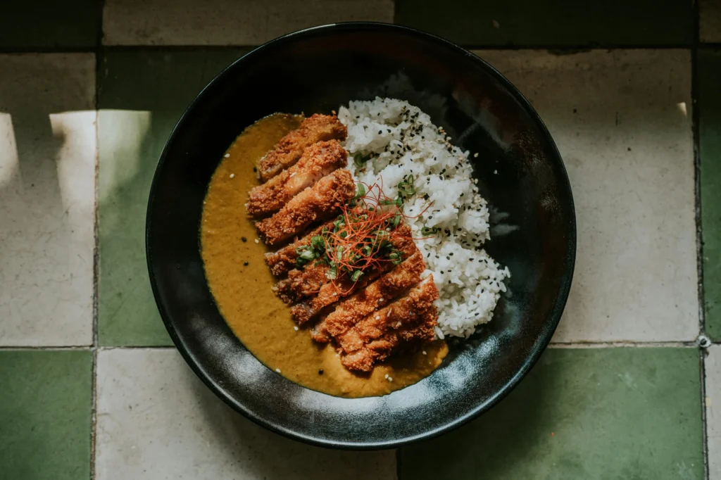 A black bowl filled with Japanese katsu curry and white rice sits centered on a green and cream tiled floor. The dish is topped with a crispy breaded cutlet, sliced into strips and garnished with green onions and fine red chili threads.