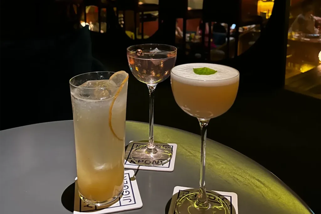 Three different artisan cocktails, including a martini and a highball, served on Jigger & Pony coasters in a dark, upscale lounge.