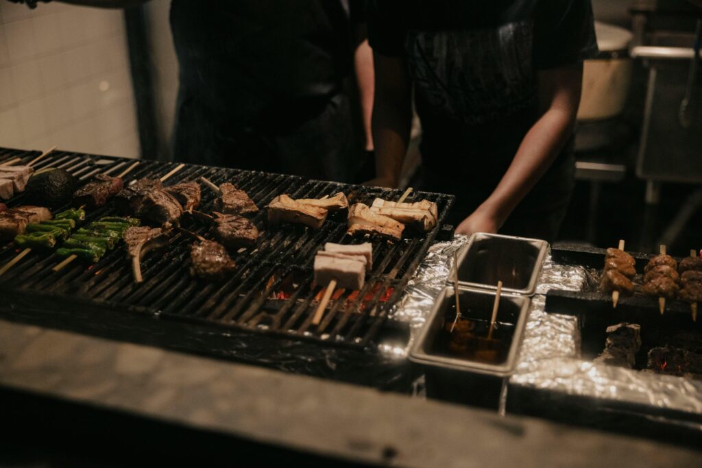 Skilled hands manage a variety of skewers, including meat, mushrooms, and peppers, as they sizzle over a glowing charcoal grill. The dimly lit kitchen setting captures the intense, focused atmosphere of traditional live-fire cooking.