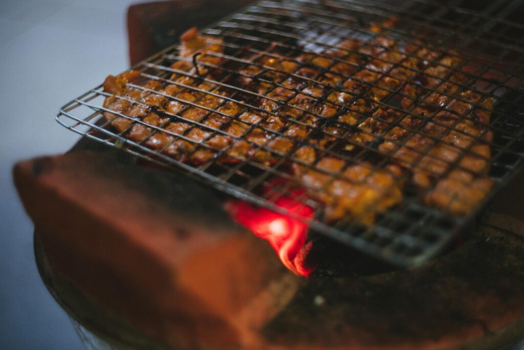 Thin strips of marinated meat are pressed within a wire grilling basket and sizzle over the glowing orange embers of a traditional clay brazier. The focused heat creates a deep char on the skewers, while a soft glow emanates from the fire beneath the rustic cooking setup.
