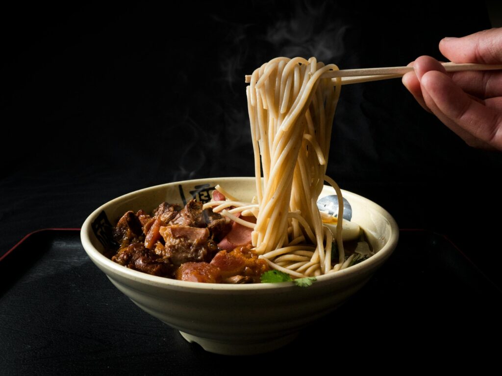 A hand uses chopsticks to lift a large swirl of steaming noodles from a ceramic bowl. The dish is filled with braised meat and a soft-boiled egg, set against a dramatic black background.