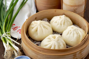 Four fluffy, steamed buns are neatly arranged inside a traditional round bamboo steamer basket. Fresh green onions tied together and stacked steamer lids in the background complete this rustic culinary scene.
