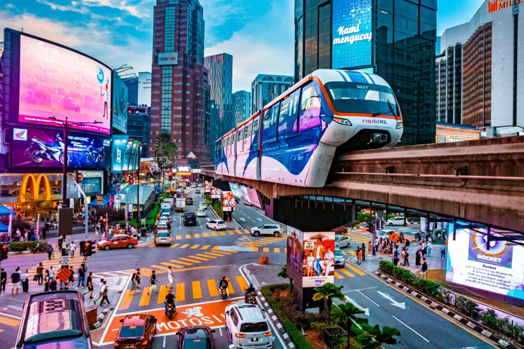 A sleek, blue and white monorail glides above a bustling neon-lit intersection in Kuala Lumpur, surrounded by vibrant billboards and skyscrapers. Below, a mix of cars, motorcycles, and pedestrians navigate the colorful crosswalks of a lively urban district.