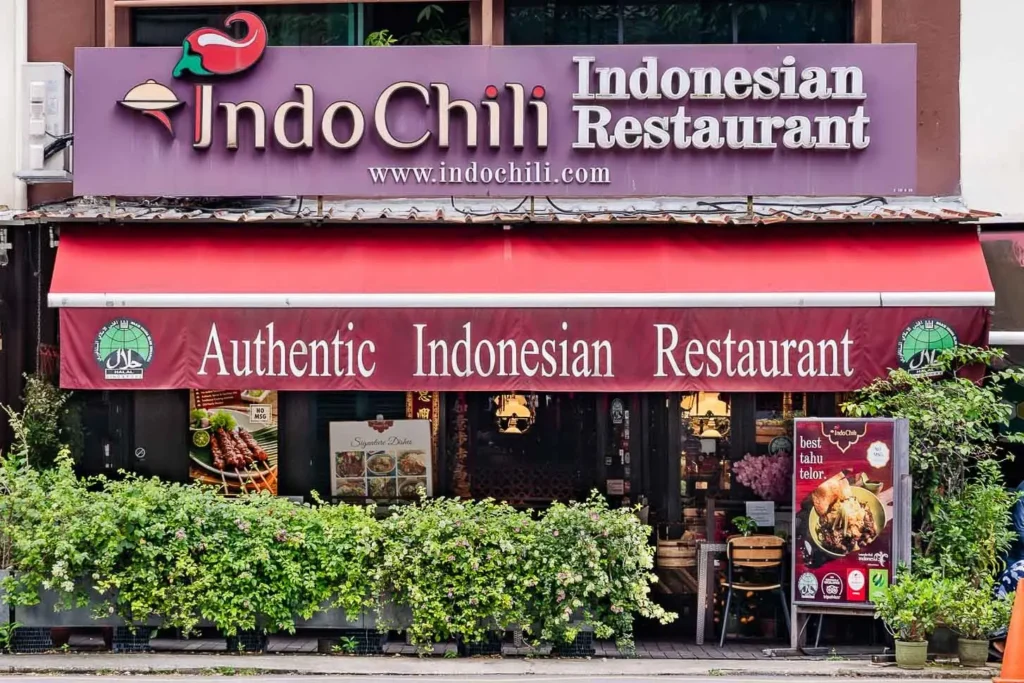 The indochili shows the exterior of a restaurant featuring a large purple sign with the name "IndoChili Indonesian Restaurant" and a prominent red awning. The entrance is lined with lush green bushes and displays signs advertising authentic Indonesian dishes and halal certification.