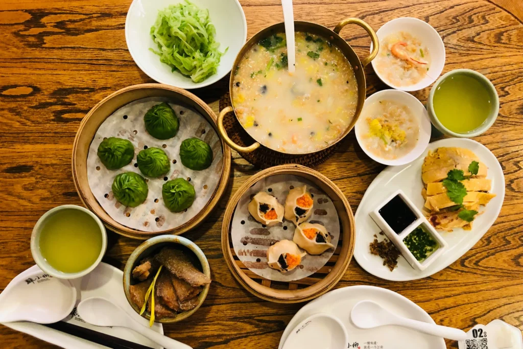 A variety of Chinese dishes, including green dumplings, siu mai, and a large pot of congee, are spread across a wooden table. The meal is completed with a plate of sliced chicken, dipping sauces, and small bowls of tea and side dishes.