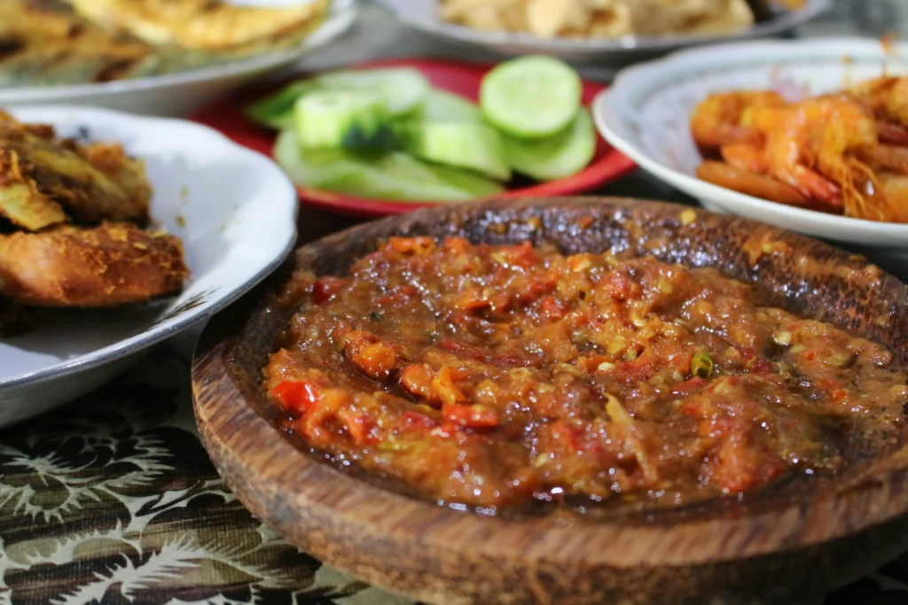 The image features a close-up of a rustic wooden bowl filled with a textured, vibrant red and orange chili paste. Surrounding the bowl are blurred plates of side dishes, including fried proteins, cooked shrimp, and sliced cucumbers on a patterned tablecloth.
