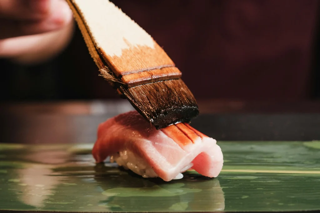 A close-up shot shows a chef using a wooden brush to apply a dark glaze or soy sauce onto a piece of tuna nigiri sushi. The sushi sits on a vibrant green leaf background, highlighting the fresh, pink marbled texture of the fish.