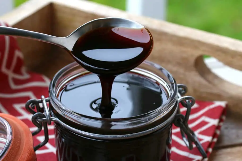 A silver spoon lifts a thick, dark brown syrup from a glass jar, showing its glossy and viscous texture. The jar sits on a wooden surface next to a red and white patterned cloth in a bright, outdoor setting.