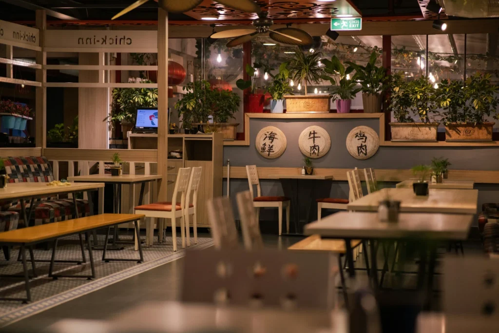 This dimly lit restaurant interior features wooden tables, patterned seating, and several potted plants arranged along a back ledge. Circular straw decorations with Chinese characters are mounted on the wall, adding a cultural touch to the warm, eclectic atmosphere.