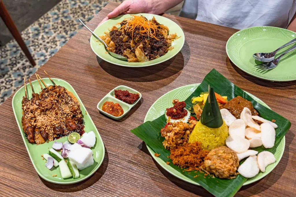 The indochili features a vibrant Indonesian feast including chicken satay with peanut sauce, a tall cone of yellow turmeric rice (Nasi Tumpeng), and a savory stir-fried dish topped with shredded carrots. The meal is presented on light green plates over a wooden table, accompanied by side dishes of sambal and fresh crackers.
