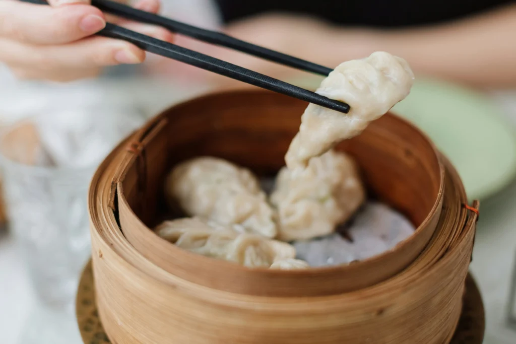 A person uses black chopsticks to lift a single steamed dumpling from a traditional bamboo steamer basket. The basket holds several other pale, pleated dumplings resting on a light-colored liner.