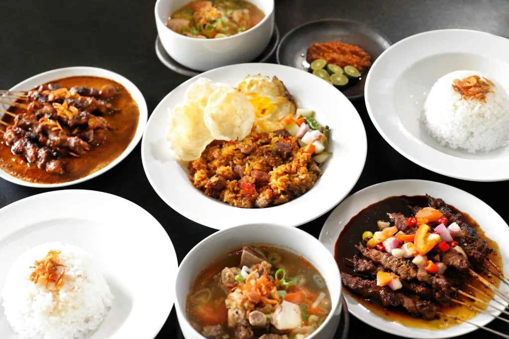 An Indonesian feast featured displays a variety of traditional dishes, including satay skewers, fried rice topped with an egg, and bowls of hearty soup. The spread is arranged on a dark table with side portions of steamed white rice, crackers, and spicy sambal.