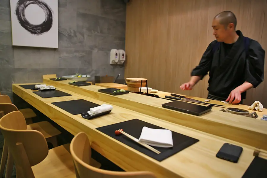 A chef prepares food behind a sleek, light wooden sushi counter set with minimalist black place settings and white linens. In the background, a large painting of a black brushstroke circle hangs on a grey textured wall next to wood-paneled accents.