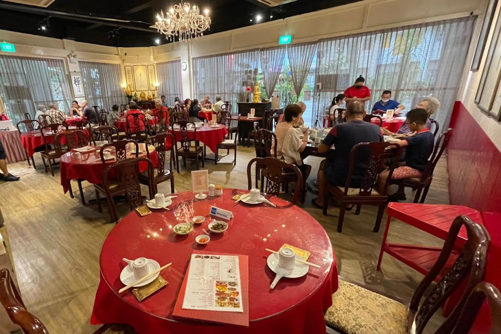 This interior shot displays a bustling Chinese restaurant featuring numerous round tables draped in vibrant red tablecloths. Patrons are seated throughout the warmly lit space, which is decorated with a crystal chandelier and traditional wooden chairs.