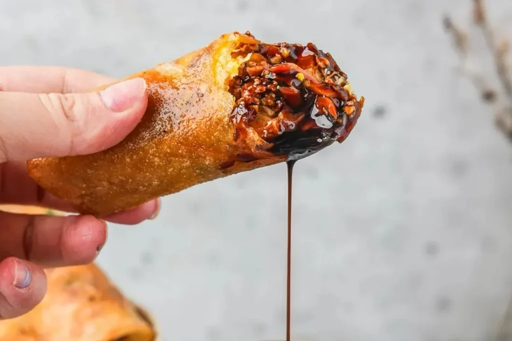 A hand holds a crispy, golden-brown spring roll that has been bitten into, revealing a savory filling of minced meat and vegetables. A thick, dark glaze—likely kecap manis—drips from the center of the roll against a neutral, textured background.