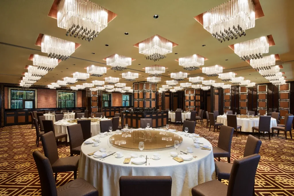 This is an elegant dining room featuring numerous round tables set with white linens and Lazy Susans. The space is illuminated by a striking grid of square crystal chandeliers and is finished with dark wood paneling and a patterned carpet.