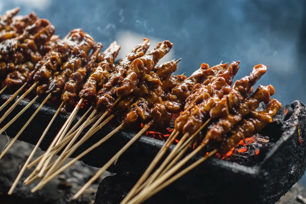 The image shows several skewers of glazed meat grilling over glowing hot coals. Wisps of smoke rise from the grill, highlighting the charred texture and rich sauce of the satay.