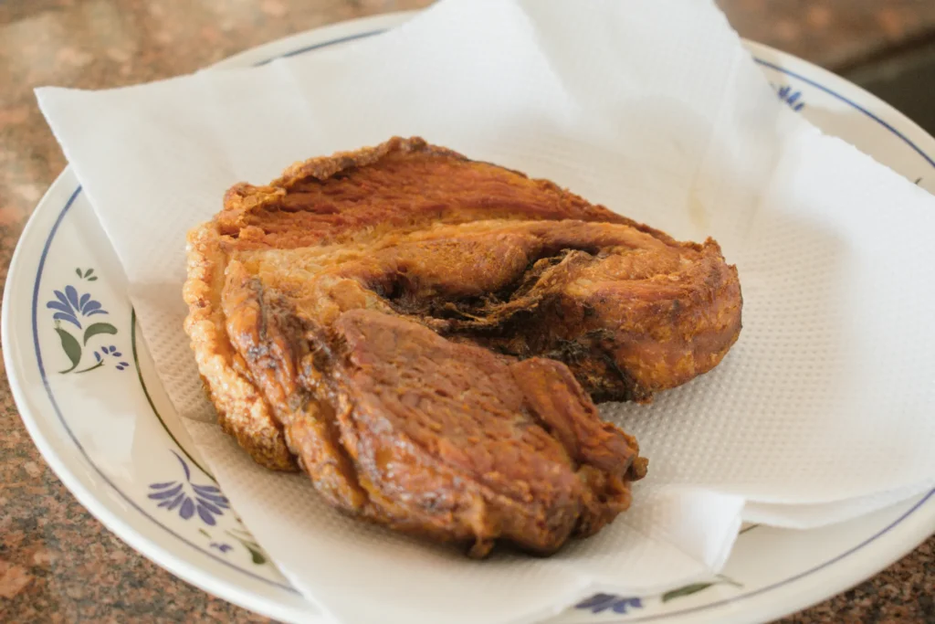 A thick, golden-brown piece of crispy fried pork belly rests on a white paper towel to drain excess oil. The meat is served on a decorative white plate featuring a delicate blue floral pattern along the rim.