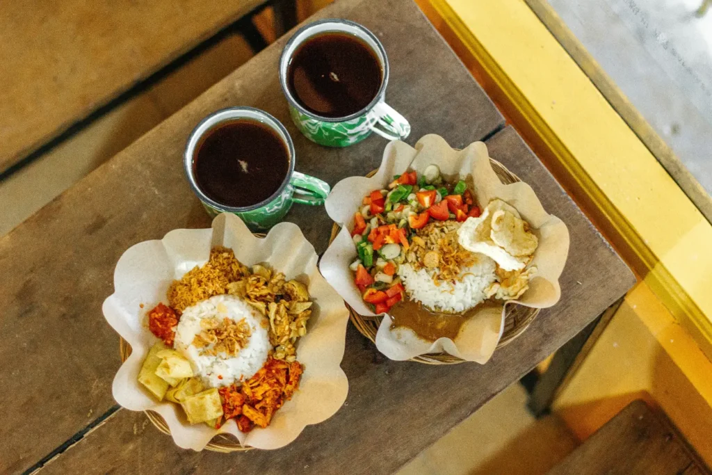 In the image, two plates of Indonesian rice dishes served on paper-lined wicker baskets sit on a rustic wooden table alongside two green patterned enamel mugs of tea. The vibrant meals feature white rice topped with fried shallots and various side dishes, including spicy sambal, fried tempeh, and fresh vegetable relish.