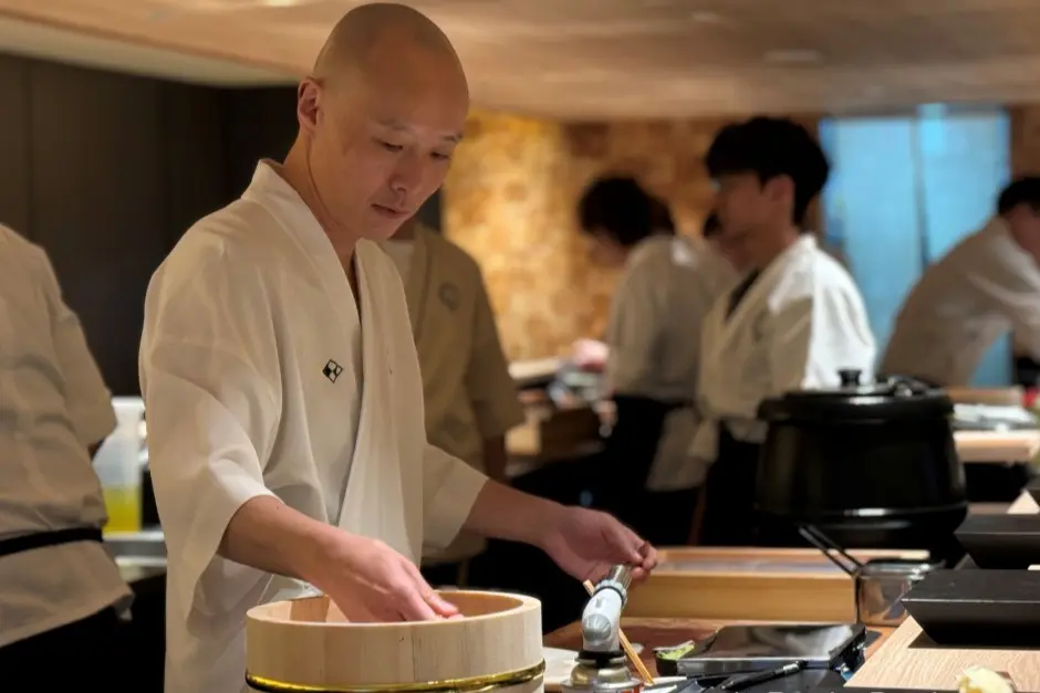 A bald Japanese chef in a traditional white uniform carefully prepares food over a wooden mixing tub at a sushi counter. In the softly lit background, other kitchen staff in matching attire are busy working in the restaurant.
