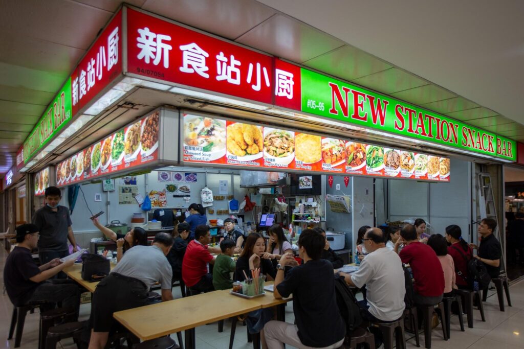 A vibrant crowd of diners is seated at long tables in front of the **New Station Snack Bar**, a brightly lit food stall featuring large, colorful menus. The scene captures the lively atmosphere of a busy indoor eatery, with staff and customers engaged under prominent red and green signage.