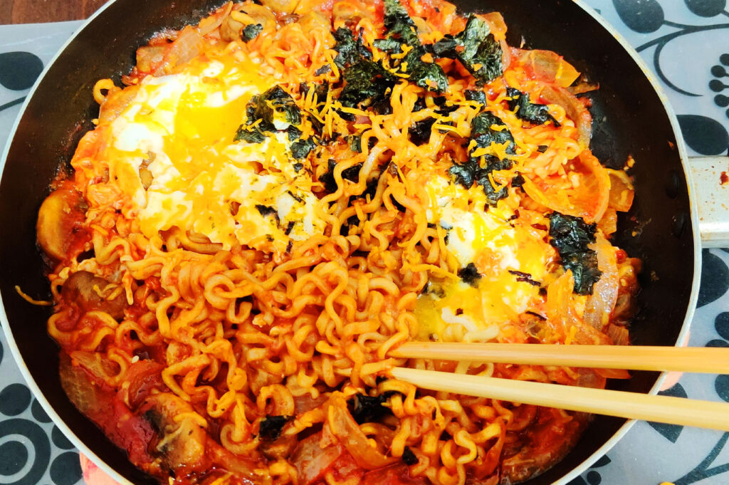Homemade Samyang spicy Korean noodles with melted cheese, egg, seaweed, and mushrooms cooked in a pan
