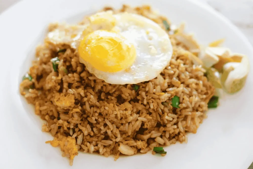 A mound of savory, golden-brown Nasi Goreng is topped with a perfectly fried sunny-side-up egg. Fresh green onions are scattered throughout the rice, which is served on a clean white plate with a side of crisp garnishes.