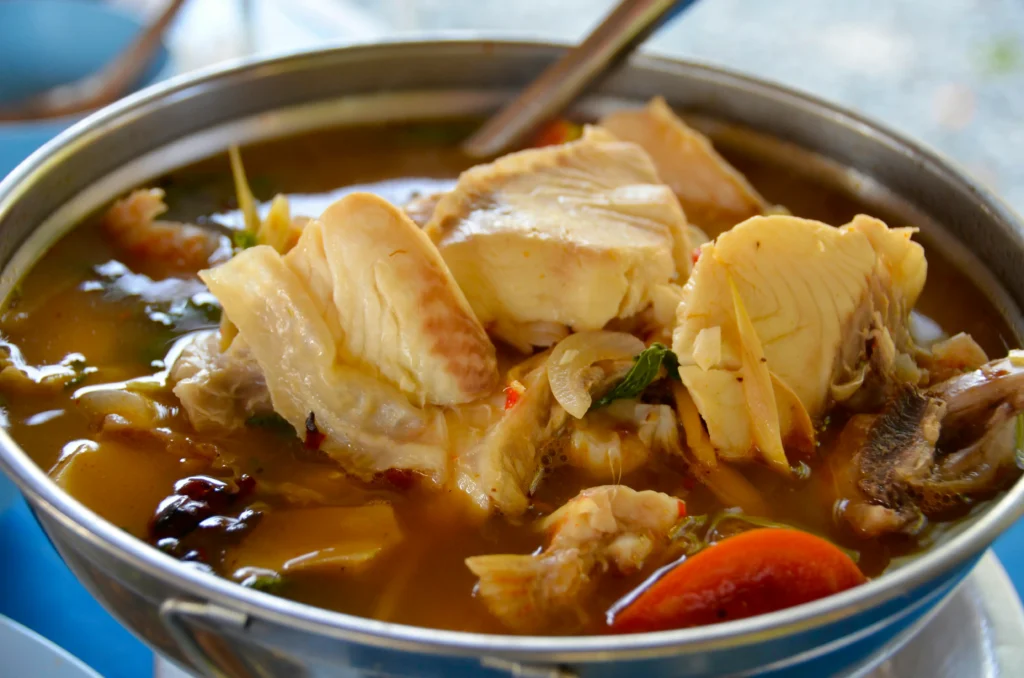 A metal pot is filled with a savory fish soup featuring large, tender chunks of white fish and sliced aromatics. The dish is served in a rich, amber-colored broth with visible ingredients like onions and tomatoes.