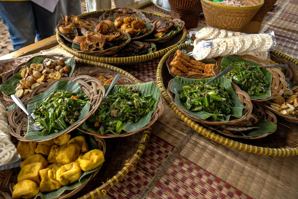 The image displays a variety of traditional Indonesian dishes served on woven platters lined with banana leaves. The spread includes stir-fried greens, fried tofu, tempeh, and crispy fish, all arranged on a checkered woven mat.