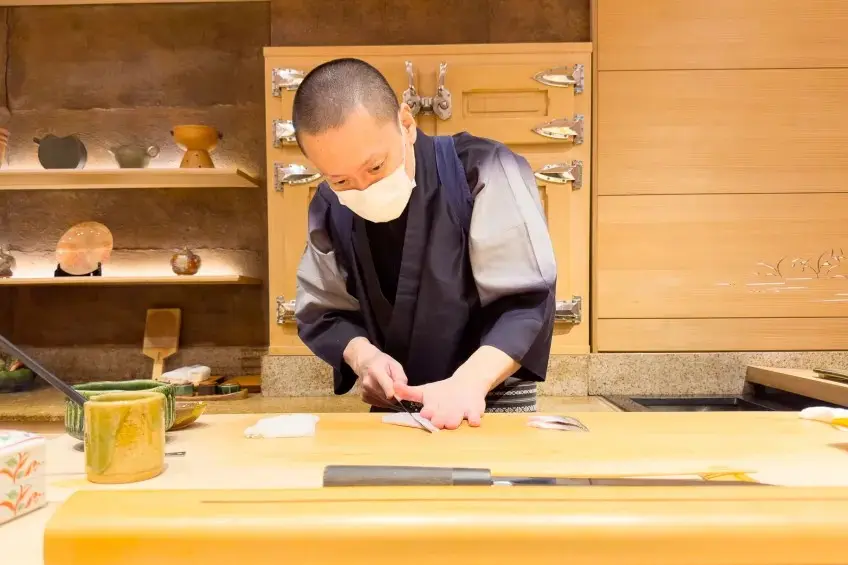 A chef wearing a face mask and traditional attire carefully slices a piece of raw fish on a wooden counter. Behind him, the upscale sushi restaurant interior features warm wooden cabinets and shelves displaying ceramic dishware.